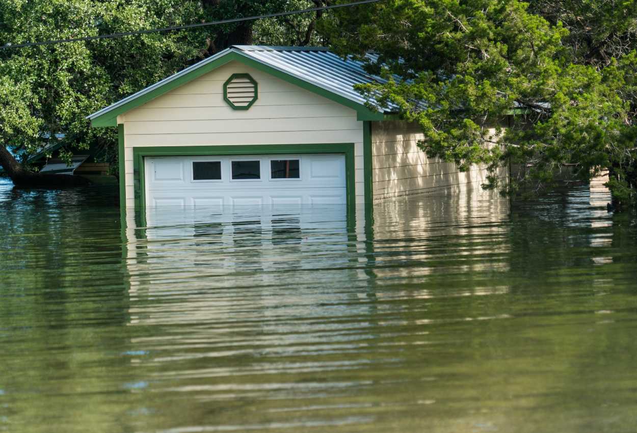 Un garage sans batardeau avec une porte blanche partiellement submergée par les eaux d'une inondation, entouré d'arbres et d'eau stagnante.